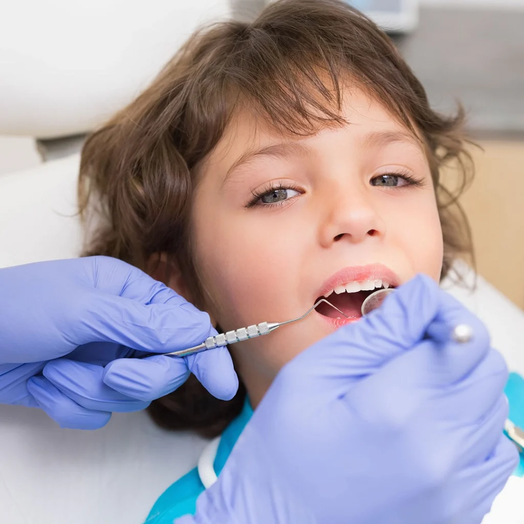 A dental professional wearing blue gloves performing an oral examination on a child seated in a dental chair, with dental instruments visible.